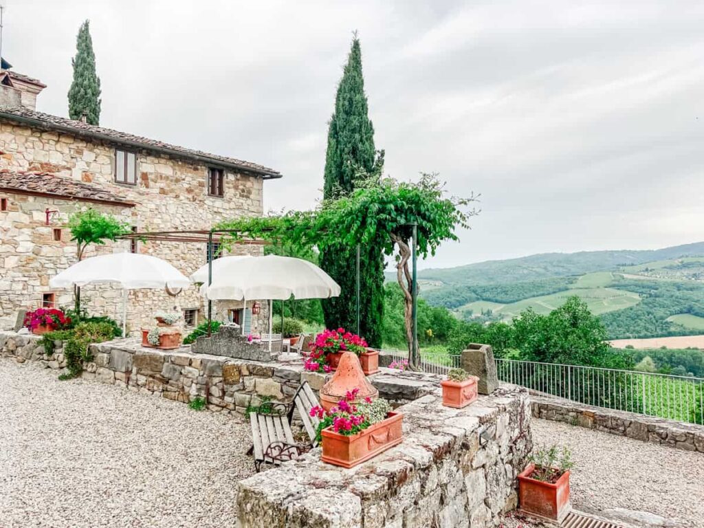 Stone farmhouse at Il Barlettaio with outdoor patio, pink flowers, white umbrellas, lush green landscape, and rolling hills in the background.