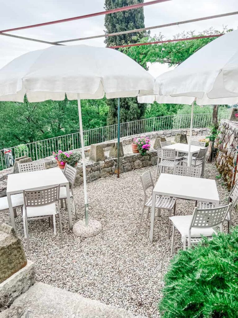 Umbrella-shaded outdoor patio with white tables and chairs, surrounded by potted pink flowers, stone retaining wall, and lush green trees in the background.