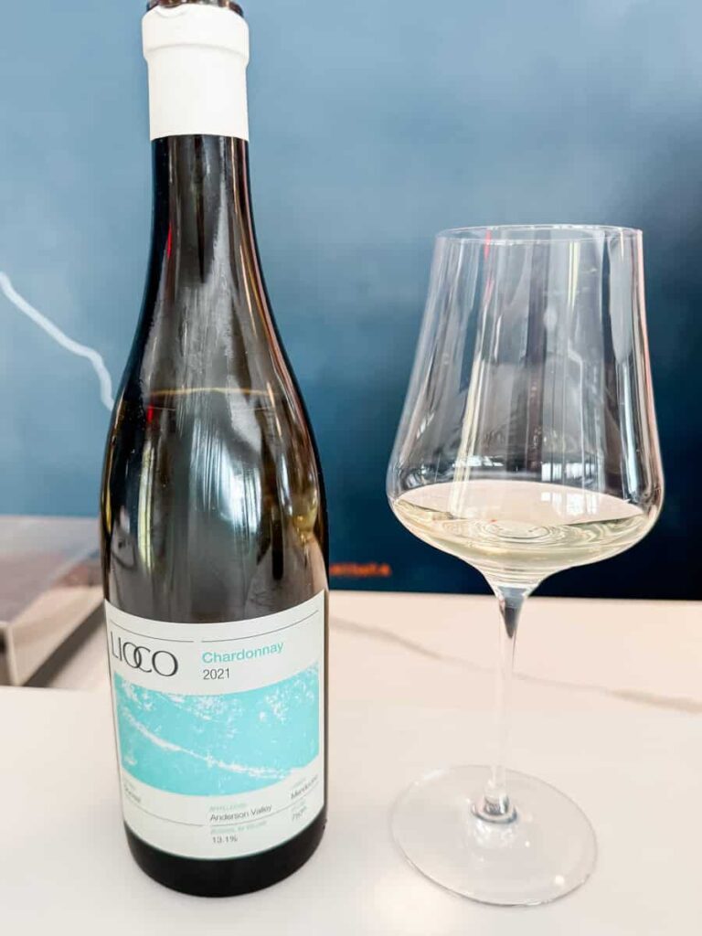 Bottle of Lioco Chardonnay wine with a white and blue label and a glass of white wine beside it on a bar surface.