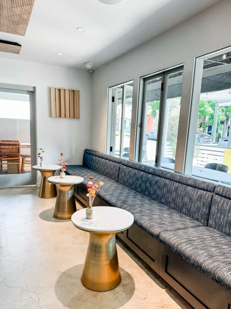 Modern lounge area at Lioco with large windows, a long upholstered bench, and small round gold and marble tables decorated with colorful flowers.