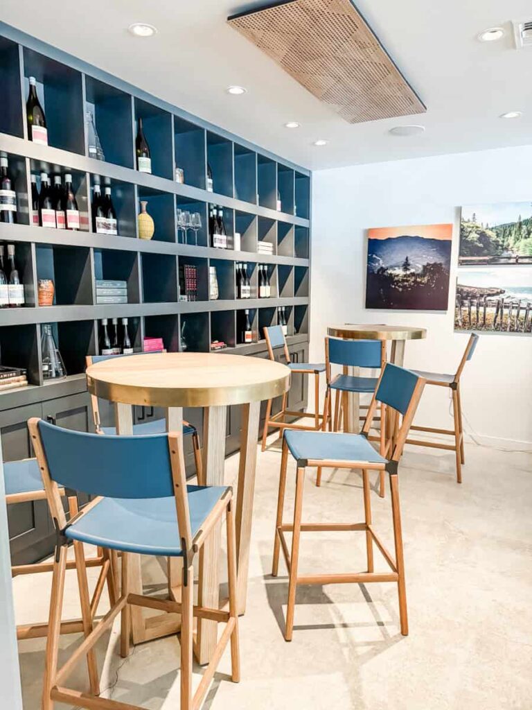 Lioco's modern wine tasting room with a large gray shelving unit filled with wine bottles, glasses, and decorative items and high-top tables with blue cushioned chairs.