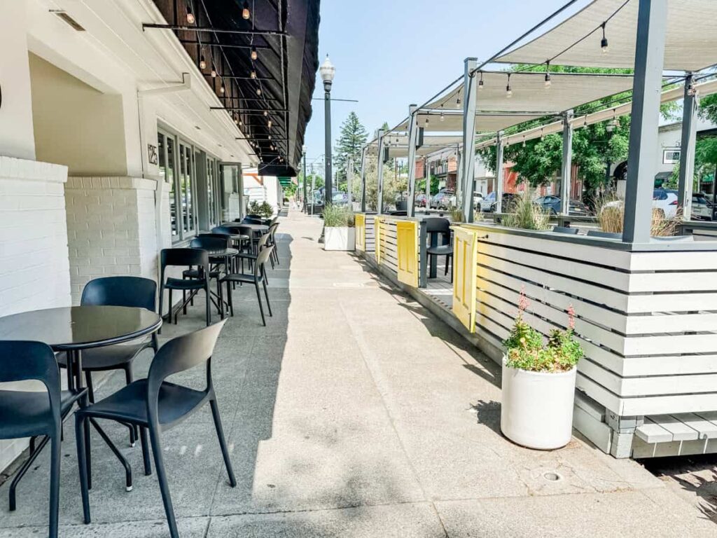 Outdoor patio at Lioco with black tables and chairs, white building wall, decorative string lights, and green plants in white pots on a sunny day.