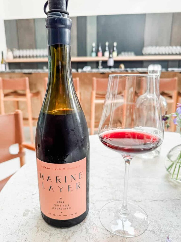 Bottle of Marine Layer Pinot Noir wine with a pink label next to a partially filled wine glass with red wine on a white marble table, and bar counter and chairs in the background.