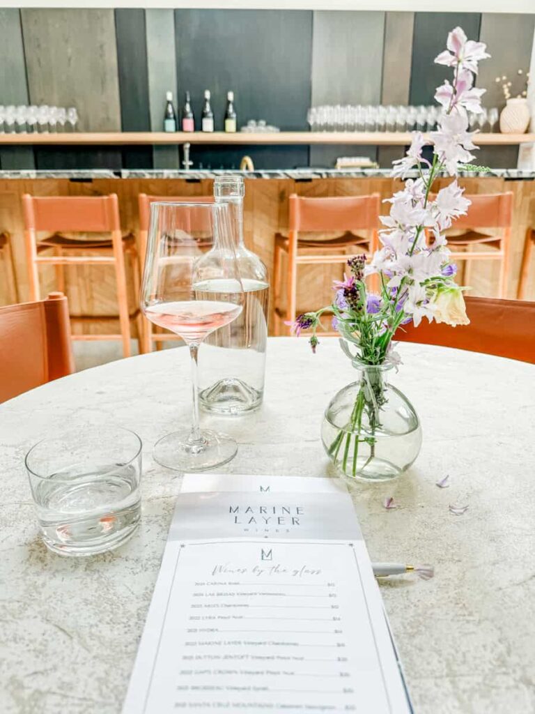 Elegant wine tasting setup at Marine Layer, featuring a wine glass, water carafe, fresh flowers in a glass vase, and a wine menu on a white table with wooden chairs in the background.