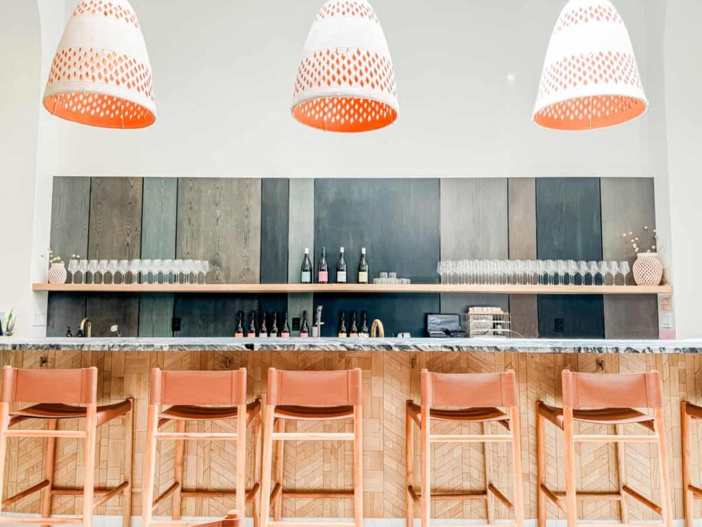 Vibrant bar interior at Marine Layer with a modern design, featuring four orange pendant lights, a marble countertop, and wooden bar stools with leather backs.