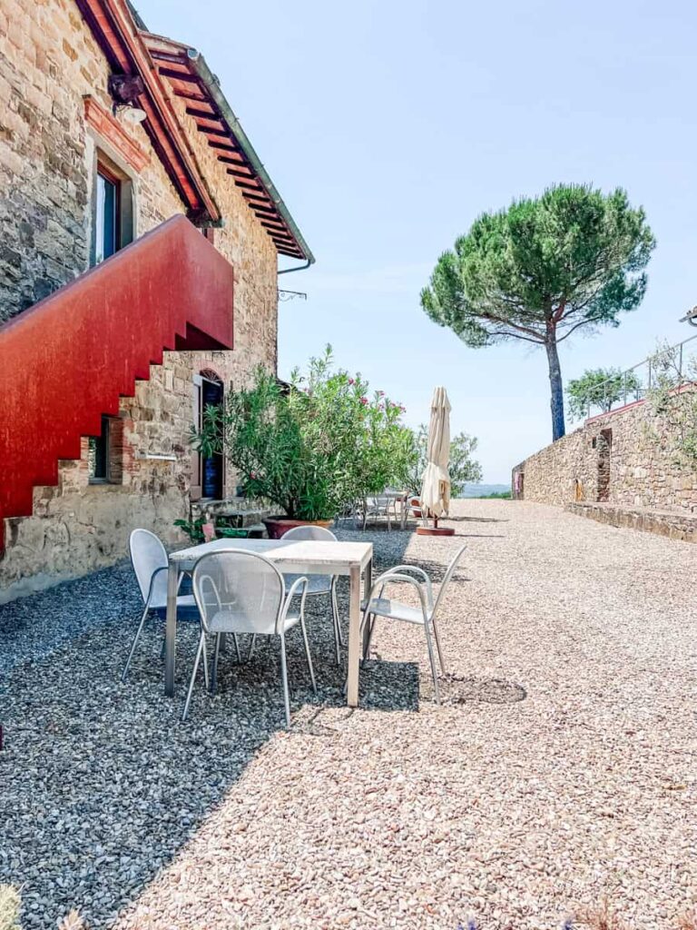 Rustic stone building with a red staircase at Poggio al Sole, surrounded by potted plants and outdoor seating on a gravel patio, with a tall green tree and clear blue sky in the background.