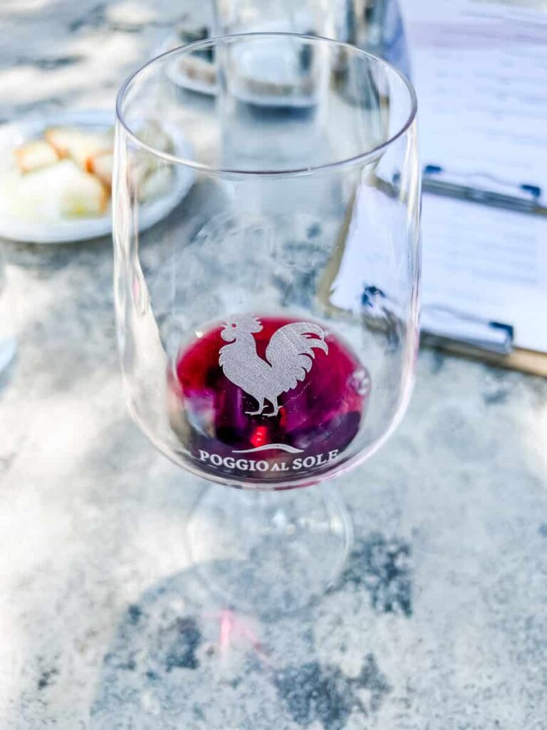 A clear wine glass with a rooster logo and the text “Poggio al Sole” contains red wine, placed on a grey marble table with a blurred background of a plate of food and a menu.