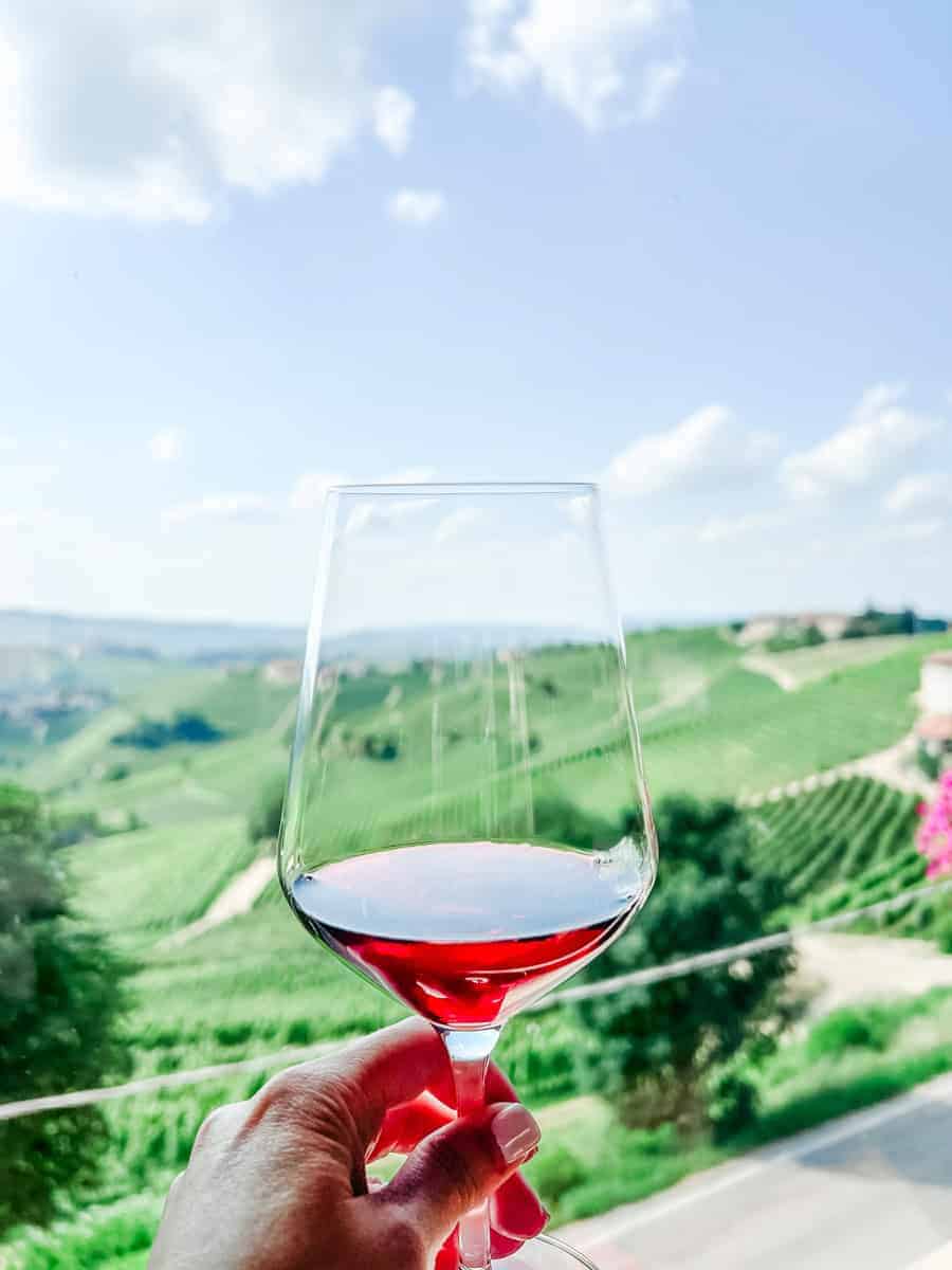 Red wine in a glass being held against a bright, cloudy sky with vibrant green rolling vineyards in the background.