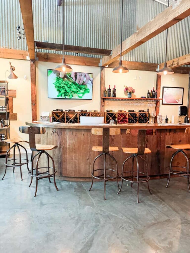 Rustic wine bar interior at Williamson Wines with wooden bar counter, high wooden chairs, wine bottles on shelves, and exposed wooden beams.