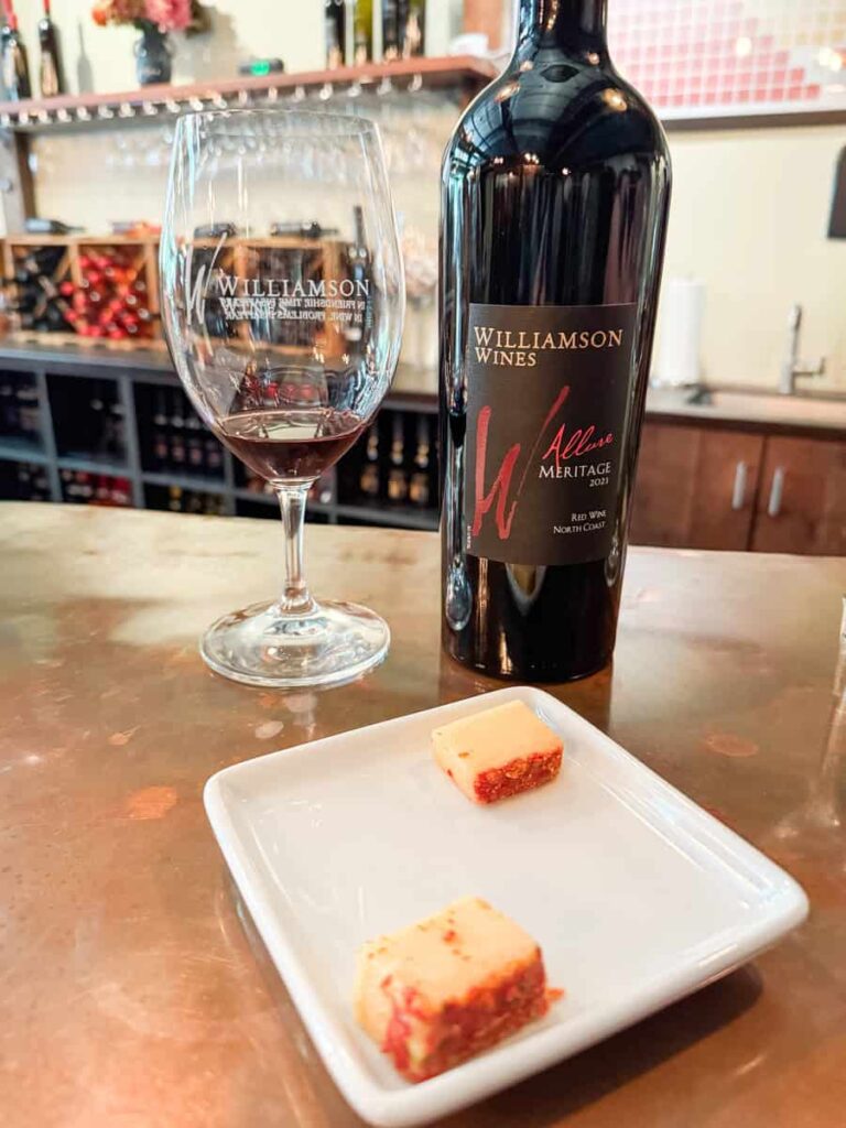 A bottle of Williamson Wines Meritage red wine, a glass of wine, and a small white plate with two cheese cubes on a copper bar top.