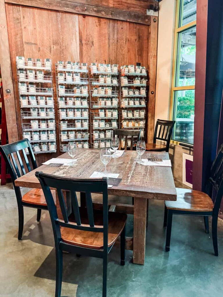 Williamson Wines tasting room with a rustic wooden table set with four wine glasses and tasting notes, surrounded by black chairs and large windows letting in natural light.
