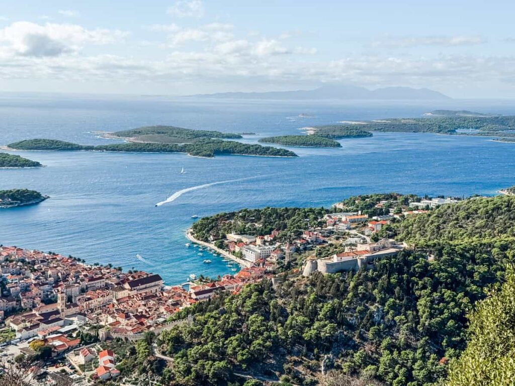Aerial view of Hvar Town with its terracotta rooftops, the hilltop Spanish Fortress, and the Pakleni Islands dotting the bright blue Adriatic Sea in the distance.