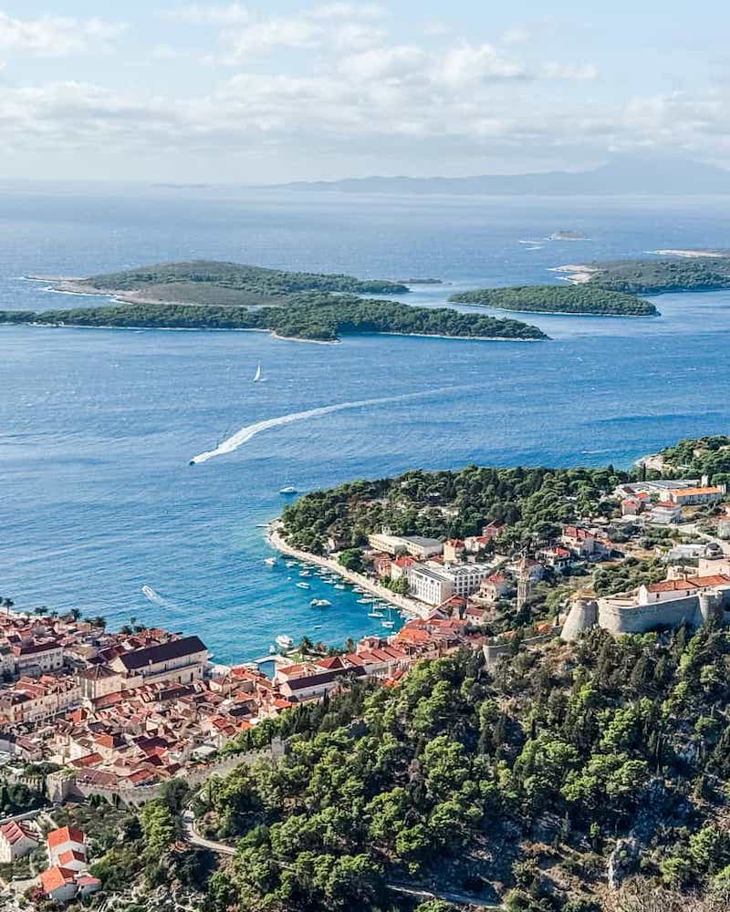 Aerial view of Hvar Town with its terracotta rooftops, the hilltop Spanish Fortress, and the Pakleni Islands dotting the bright blue Adriatic Sea in the distance.
