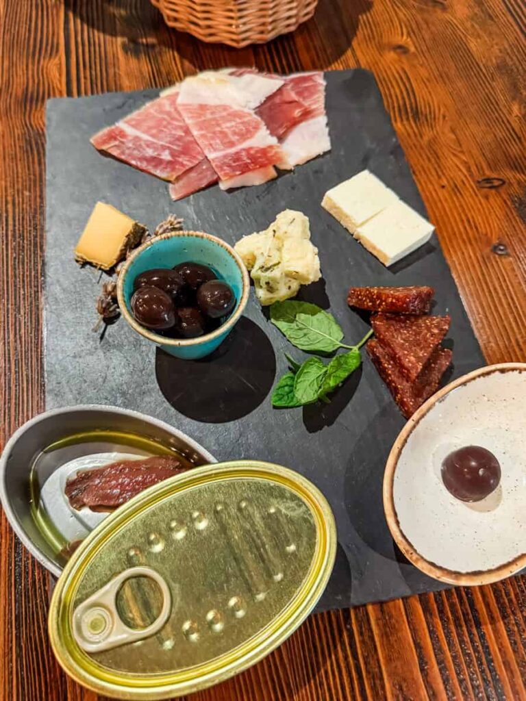 Charcuterie board at Vina Carić featuring slices of cured meat, assorted cheeses, black olives, a tin of anchovies in olive oil, and dried fig pieces, served on a slate platter with rustic wooden table beneath.