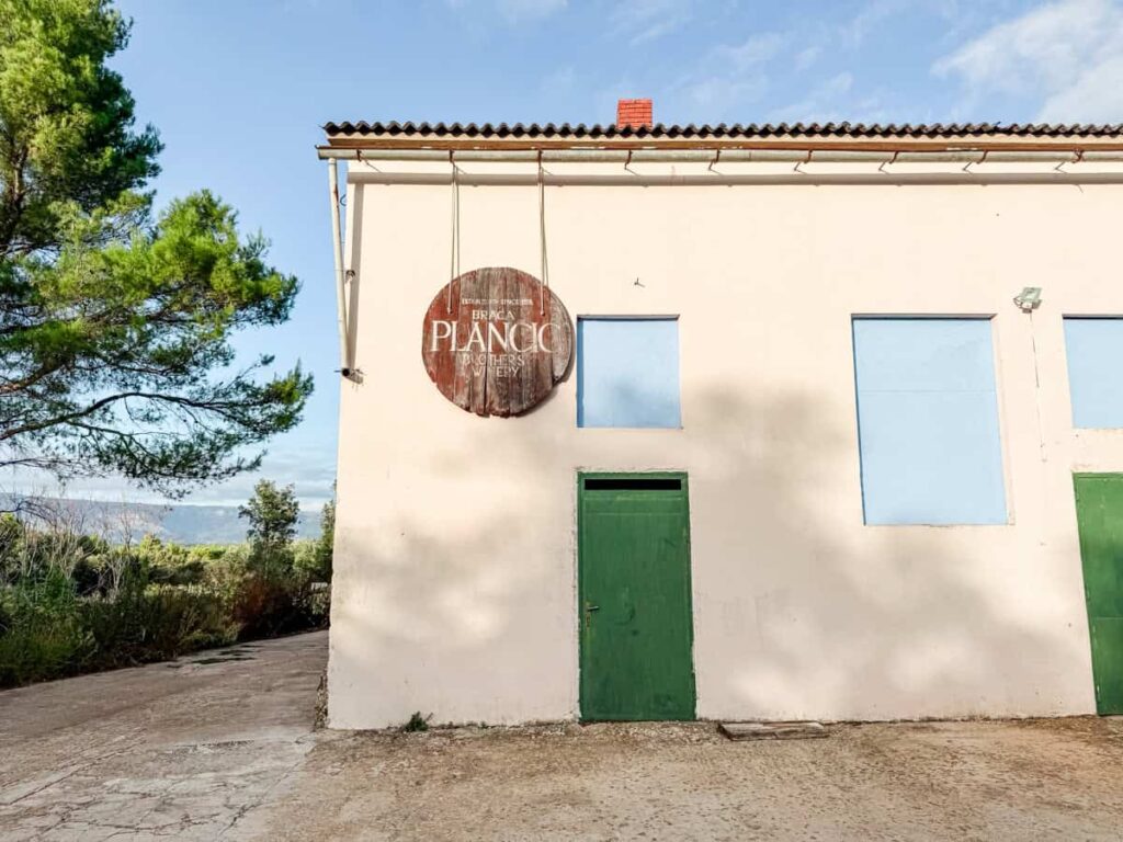 Exterior of Braća Plančić Brothers Winery, with a wooden barrel sign mounted above a green door on a pale stucco building.