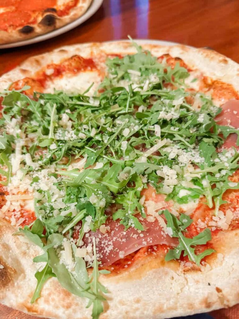 Close-up of a wood-fired pizza with tomato sauce, prosciutto, fresh arugula, and shaved cheese, served on a wooden table.