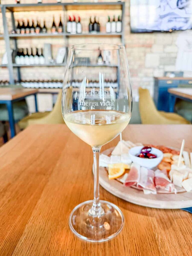 Glass of white wine at Merga Victa Winery tasting room with charcuterie board in background.
