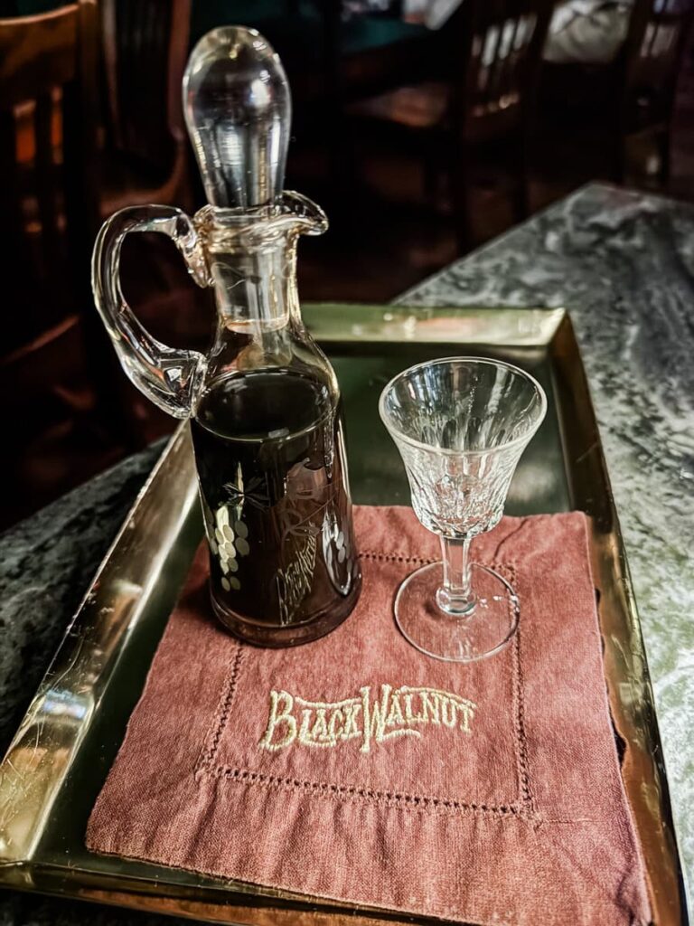 A small etched glass carafe filled with dark liqueur and a cordial glass served on a tray with a brown napkin embroidered with the Black Walnut logo.