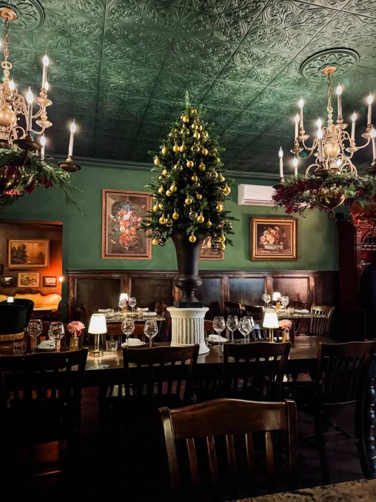 Elegant interior of Black Walnut restaurant featuring deep green walls, chandeliers, framed paintings, and a central table set for dinner beneath a Christmas tree decorated with gold ornaments.