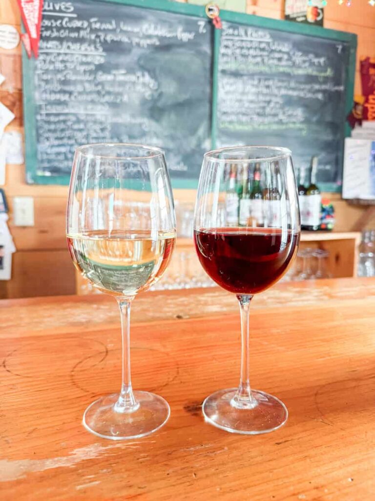 Close-up of a glass of Nebbiolo wine and a glass of Grillo wine on a wooden bar, with blurred chalkboard menus in the background.