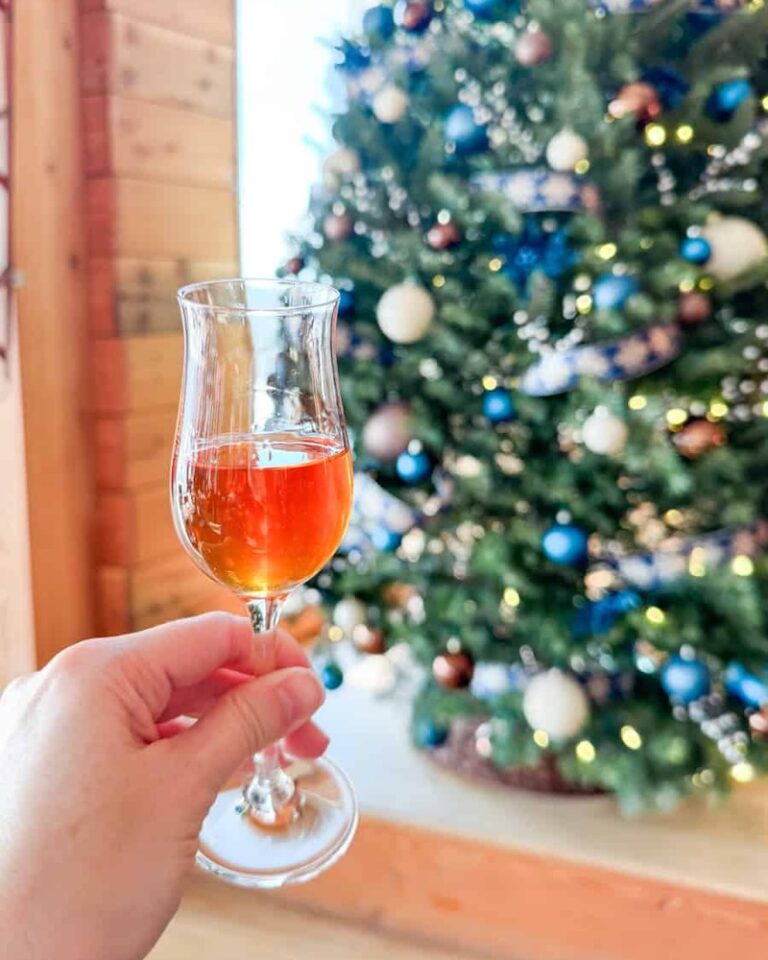 Hand holding a glass of amber-colored Seminary Hill cider in front of a Christmas tree decorated with blue, white, and brown ornaments.
