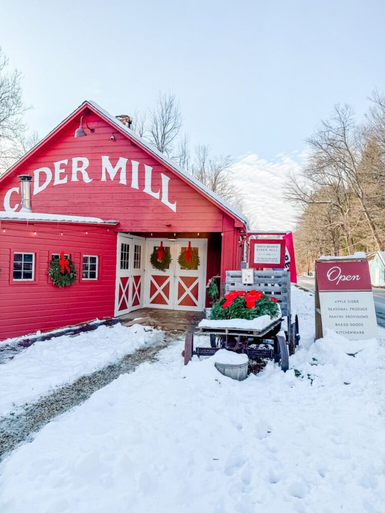 Bright red barn with “Cider Mill” painted across the top, festive wreaths on the doors, and a sign listing apple cider, produce, and baked goods, surrounded by fresh snow.