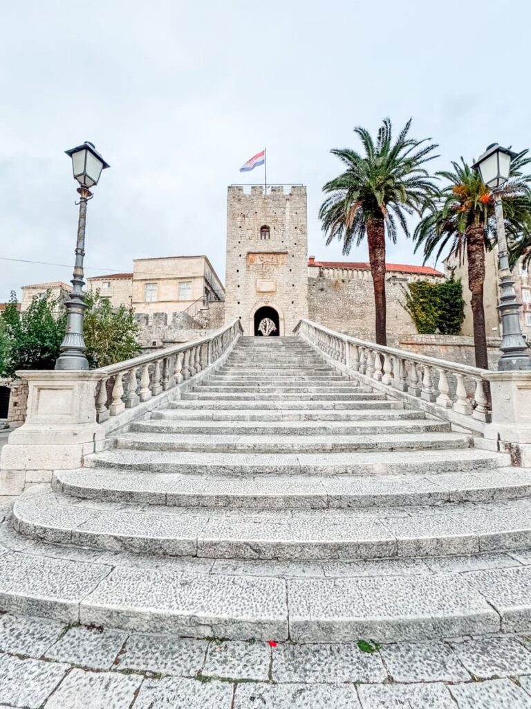 Stone staircase leading to a historic gate tower in Korčula Old Town with palm trees and Croatian flag.