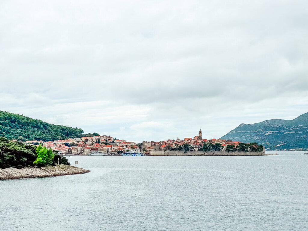 Panoramic view of Korčula Old Town from across the water, with stone walls, red-roofed buildings, and mountains in the background.