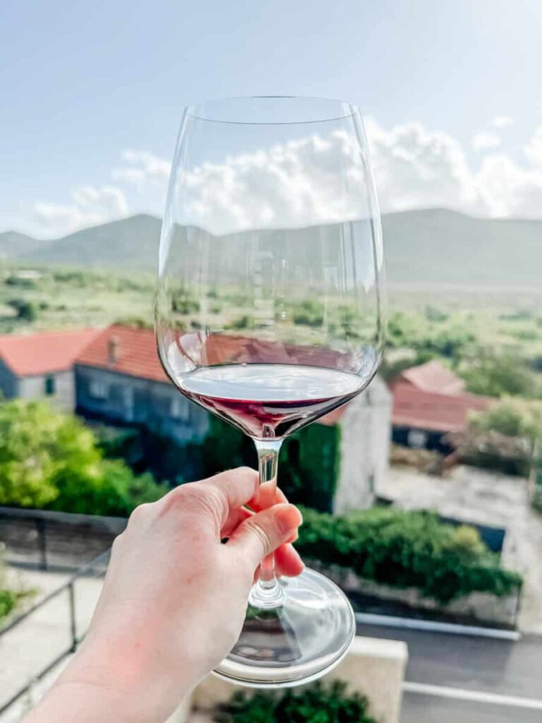 Glass of red wine held up on a balcony at Miloš Winery with vineyard-covered hills in the background.