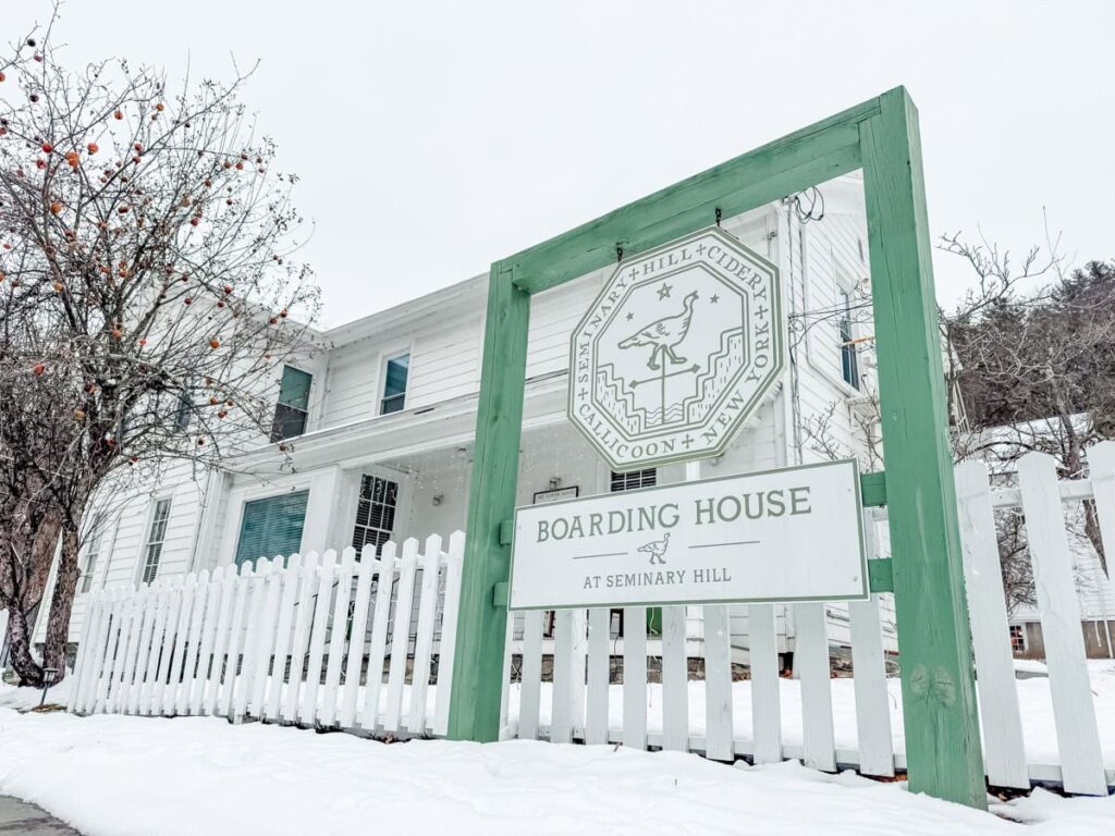 White farmhouse with a picket fence and green Seminary Hill Boarding House signage, surrounded by snow and a bare apple tree.