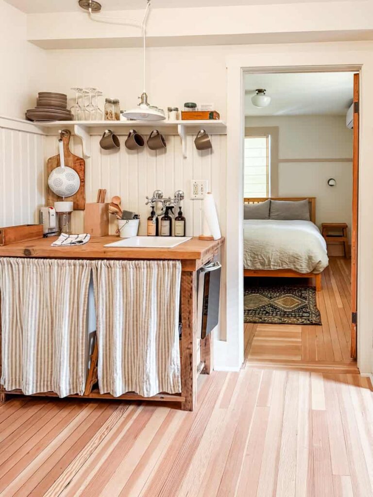 Boarding House in-room kitchenette with rustic wood details, shelves of dishware, and a view into the bedroom with a neatly made bed.