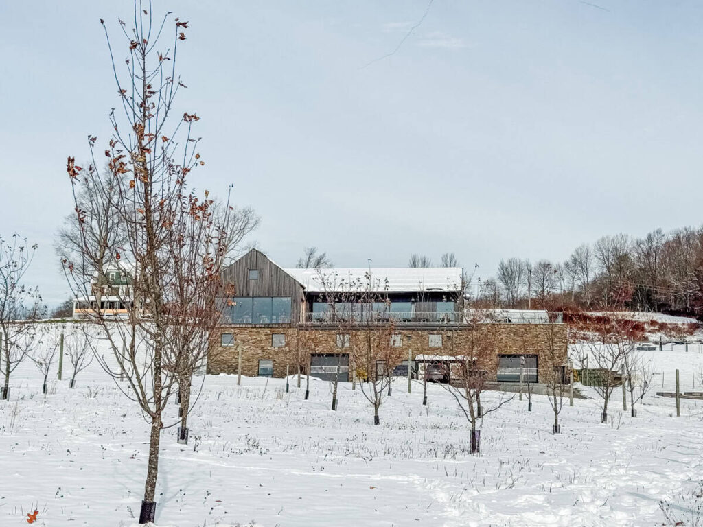 Modern cidery building surrounded by snowy orchards, blending wood and stone architecture with large windows.
