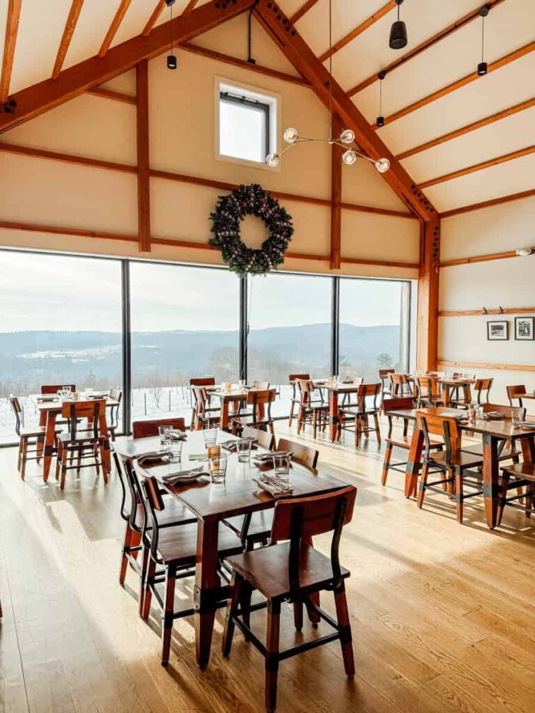 Sunlit tasting room with wood-beamed ceilings, a large wreath, and panoramic windows overlooking snow-covered orchards.