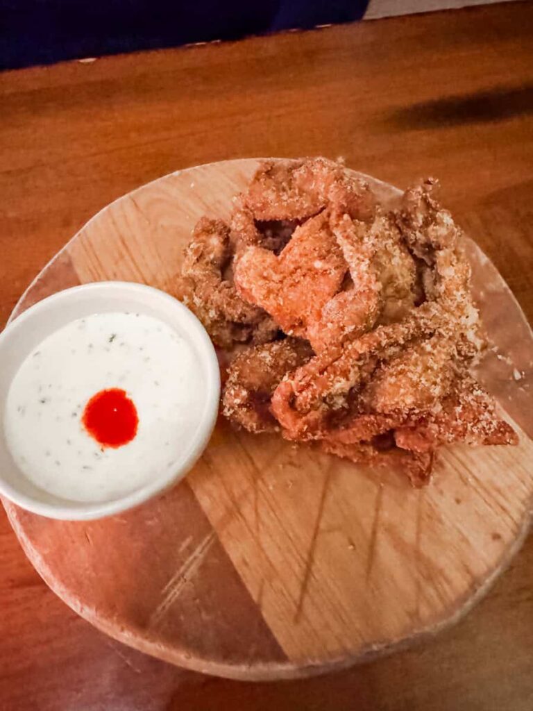 Plate of crispy fried chicken skins served on a wooden board with a side of creamy dipping sauce topped with a dot of hot sauce.