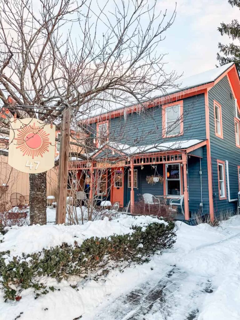 Snow-covered path leading to the dark blue and coral-trimmed Sunshine Colony wine bar, with a wooden sign reading “47” hanging from a tree.