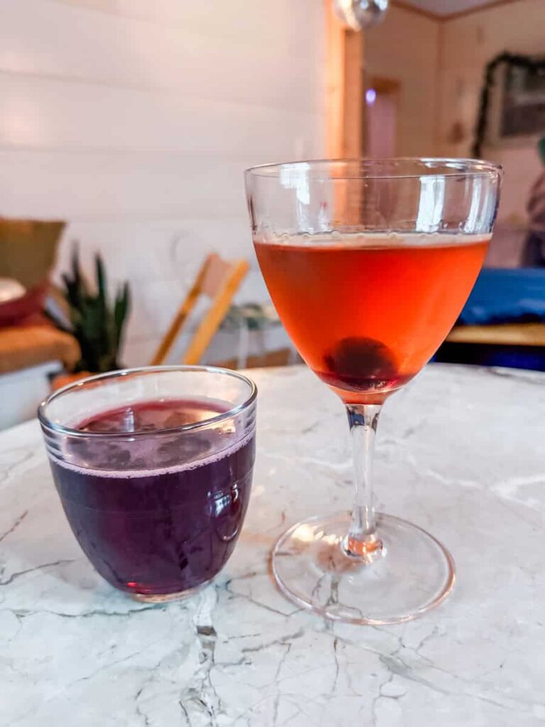 A clear coupe glass with an Manhattan cocktail and a short glass with Lambrusco wine, both resting on a marble table.