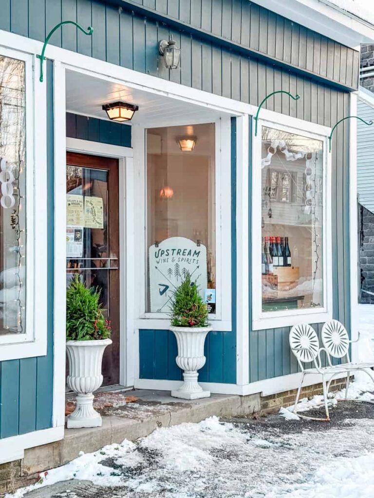 Blue and white exterior of Upstream Wine & Spirits with a glass door and window signage, flanked by potted evergreens and surrounded by snow.