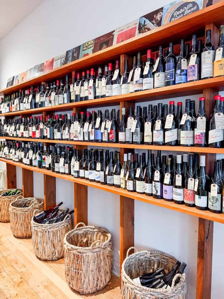 nterior of Upstream Wine & Spirits featuring wooden shelves lined with wine bottles labeled with handwritten tags, with vinyl record covers displayed above and wicker baskets below.