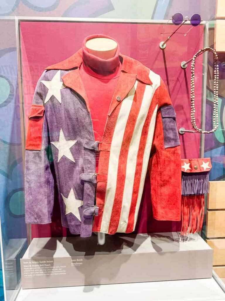 Patriotic suede jacket in red, white, and blue with stars and stripes, displayed with vintage sunglasses and a beaded necklace at the Bethel Woods museum.