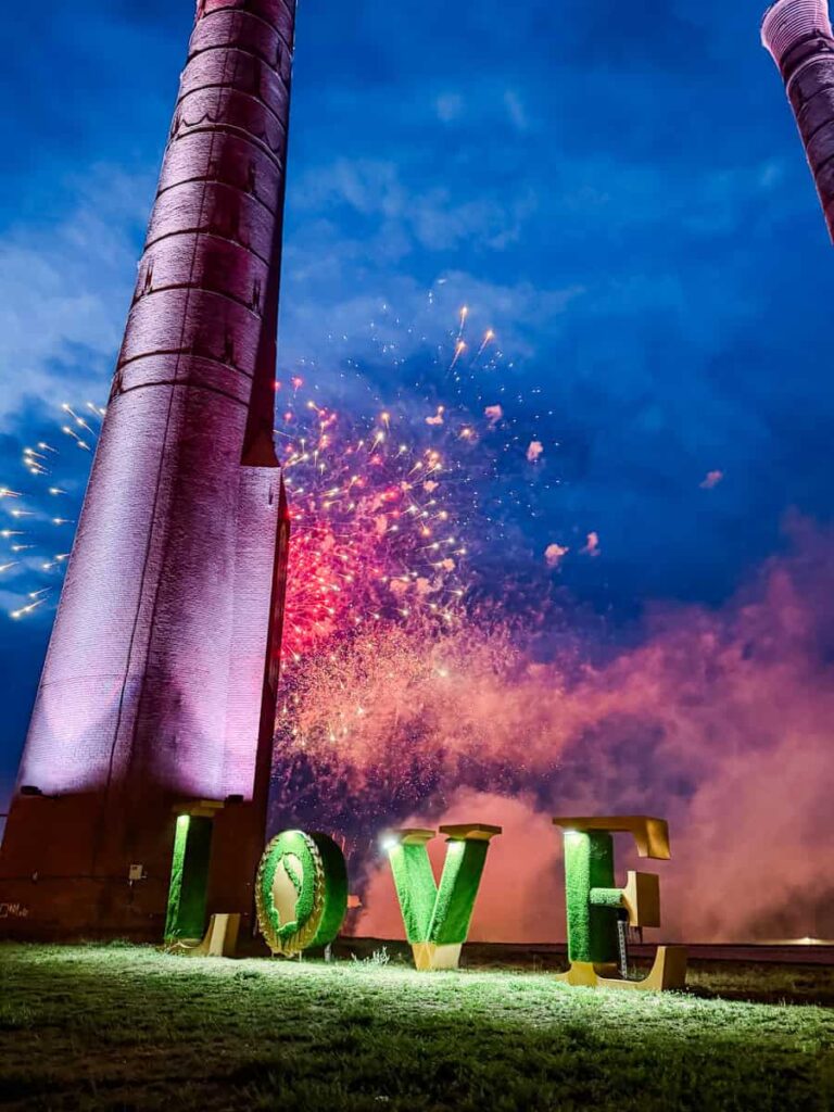 Fireworks burst behind the illuminated LOVE sculpture and historic smokestacks outside Caesars Virginia at dusk.