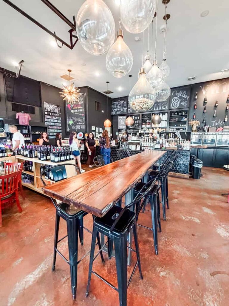 Interior of Crema & Vine with a long communal table, hanging glass pendant lights, wine displays, a self-pour wine station, and a coffee bar.