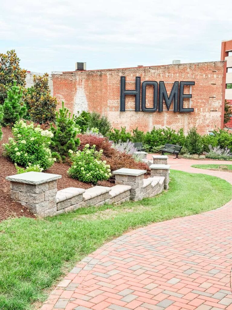 Landscaped park in Danville's River District with winding brick paths, garden beds, benches, and a large black "HOME" sign mounted on a brick wall.