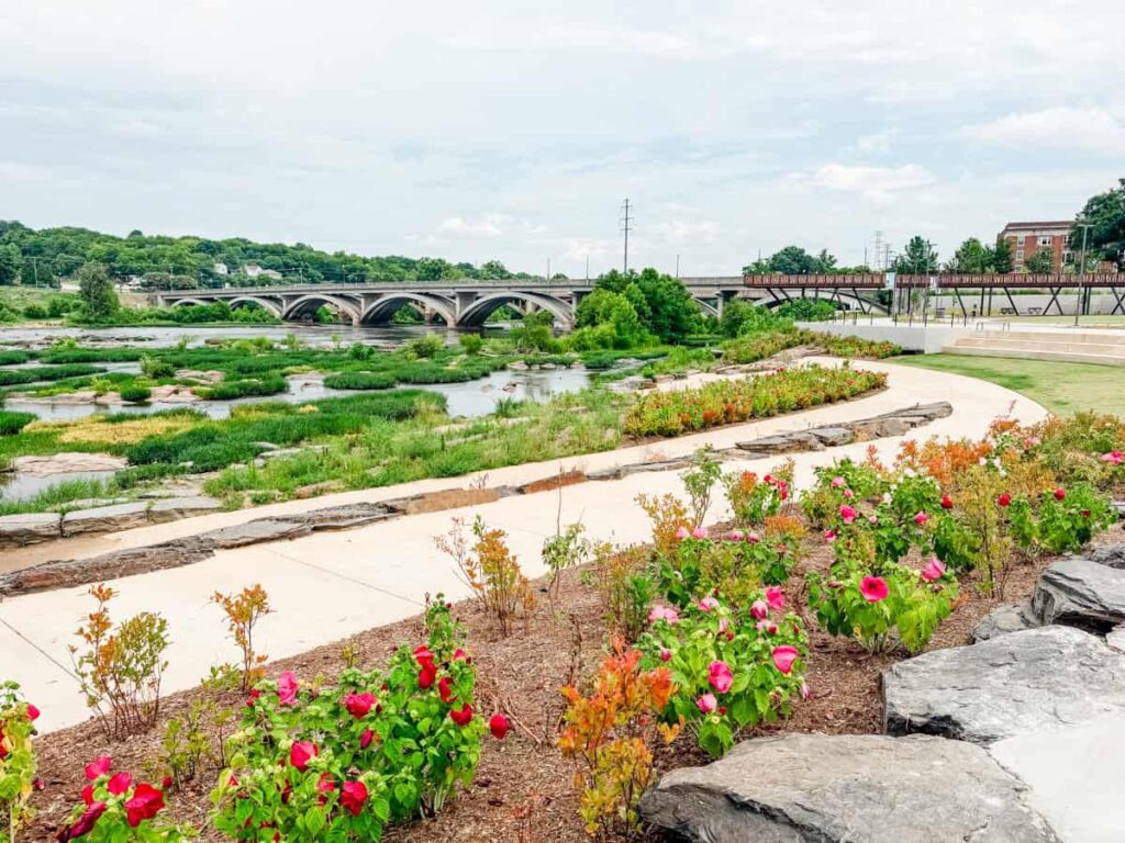 Curving walking paths and colorful flower beds line Danville Riverfront Park beside the Dan River, with rocky river islands, a pedestrian bridge, and an arched bridge creating a scenic view of the River District waterfront.