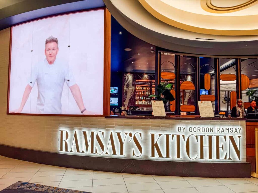 Entrance to Ramsay's Kitchen featuring a large illuminated sign reading "Ramsay's Kitchen by Gordon Ramsay" and a portrait of Gordon Ramsay above the host stand.