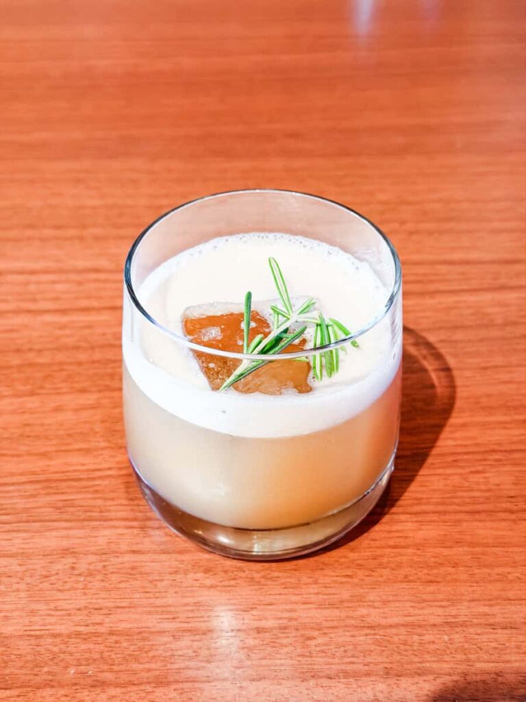 Craft cocktail with egg white foam garnished with a rosemary sprig from a mixology demonstration at Ramsay's Kitchen.