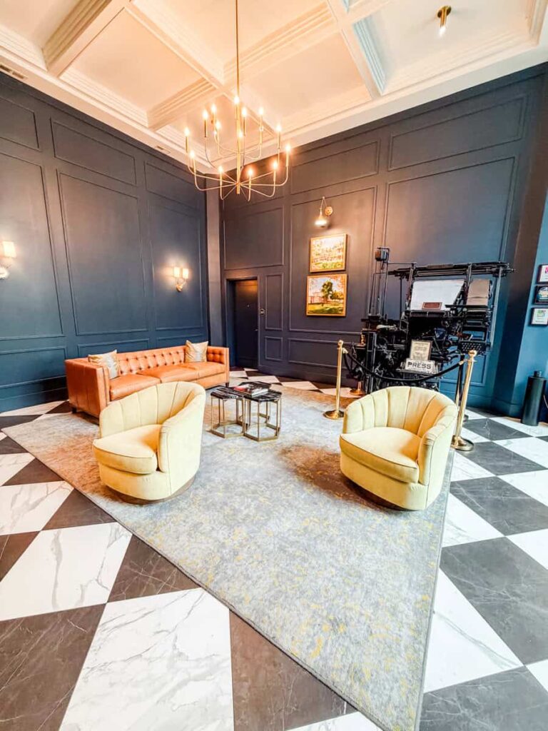 Hotel lobby at The Bee with black paneled walls, leather seating, a chandelier, and a historic printing press on display.