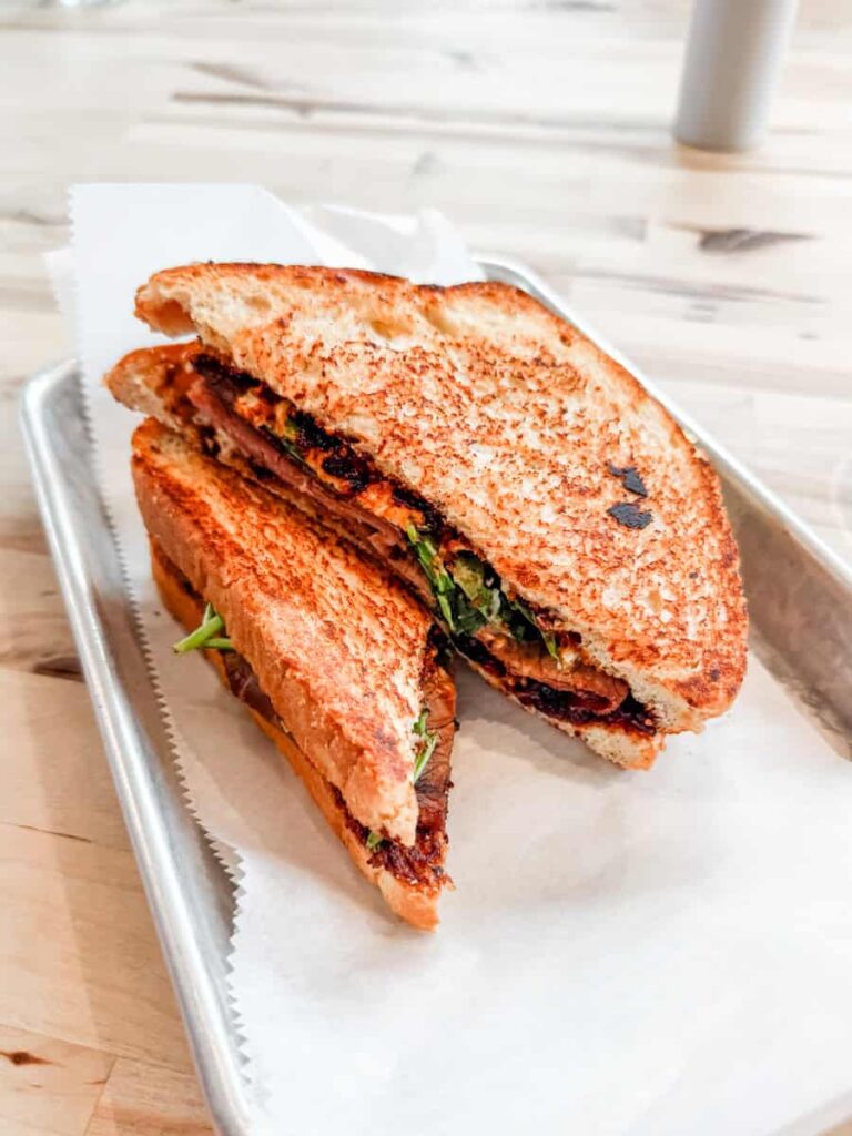 Toasted sandwich filled with roast beef, fig spread, spinach, and goat cheese cut in half and served on a metal tray with parchment paper at The Pod restaurant in Gretna.