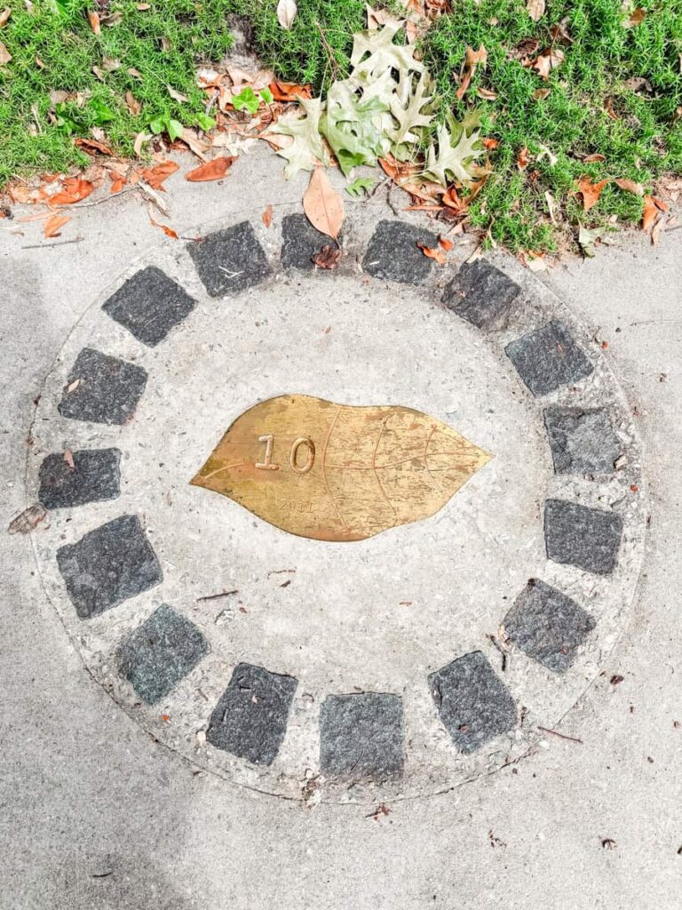 Brass tobacco leaf marker embedded in the sidewalk, surrounded by stone pavers and grass. The marker is engraved with the number "10."